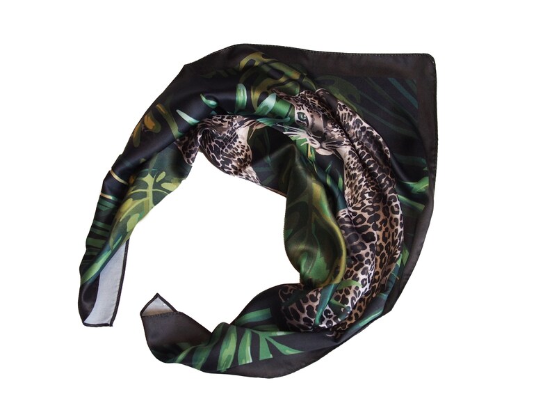 Op de afbeelding: Een zwarte zijden sjaal met een jungledesign. De sjaal heeft een luipaardprint en groene tropische bladeren. De sjaal is opgevouwen en toont de gedetailleerde print en kleurvariaties.