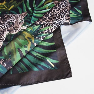 Op de afbeelding: Een vierkante zijden sjaal met een luipaardprint en junglethema. De sjaal heeft een zwarte rand en een gedetailleerde illustratie van een luipaard te midden van weelderig groen gebladerte. De sjaal is opgevouwen en onthult de onderkant.