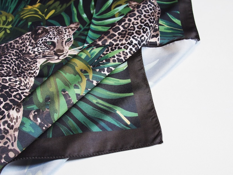Op de afbeelding: Een vierkante zijden sjaal met een luipaardprint en junglethema. De sjaal heeft een zwarte rand en een gedetailleerde illustratie van een luipaard te midden van weelderig groen gebladerte. De sjaal is opgevouwen en onthult de onderkant.