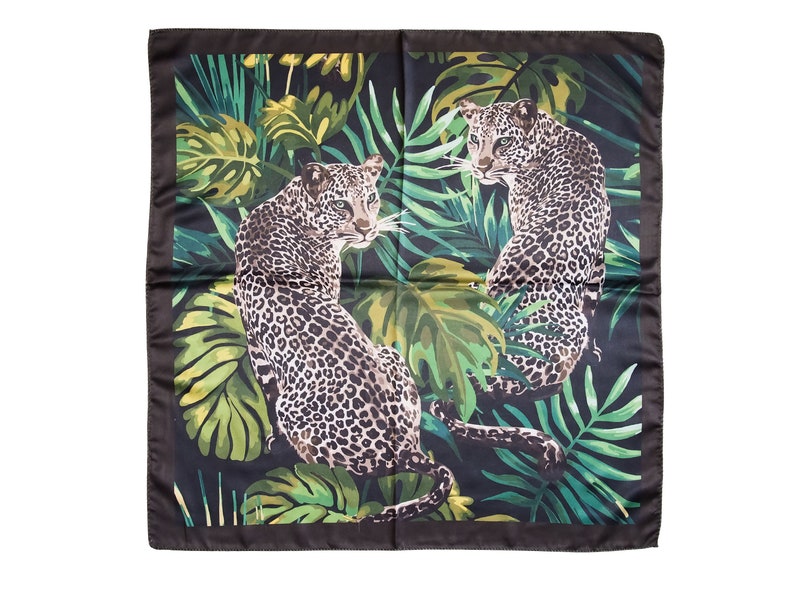 Op de afbeelding: Vierkante zwarte sjaal met een junglethema. Twee luipaarden met gevlekte vacht staan tegen een achtergrond van weelderig groen gebladerte. De sjaal heeft een zwarte rand en een satijnen afwerking.