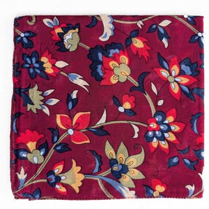 May include: A square, burgundy scarf with a floral pattern. The design features stylized flowers in shades of red, yellow, blue, and green, set against a deep red background. The scarf is likely made of a soft fabric.