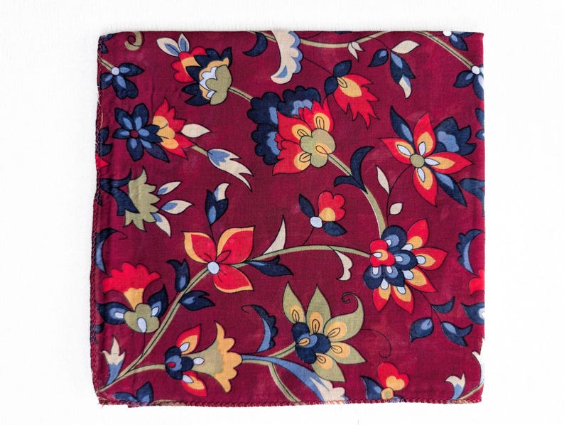 May include: A square, burgundy scarf with a floral pattern. The design features stylized flowers in shades of red, yellow, blue, and green, set against a deep red background. The scarf is likely made of a soft fabric.