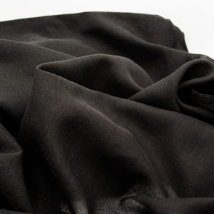 May include: Close-up of a black fabric with soft folds and a slightly textured surface. The fabric appears to be lightweight and drapes naturally, creating shadows and highlights. The overall impression is of a dark, flowing material.