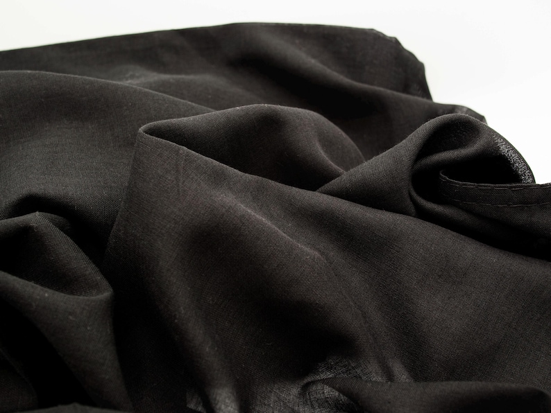 May include: Close-up of a black fabric with soft folds and a slightly textured surface. The fabric appears to be lightweight and drapes naturally, creating shadows and highlights. The overall impression is of a dark, flowing material.