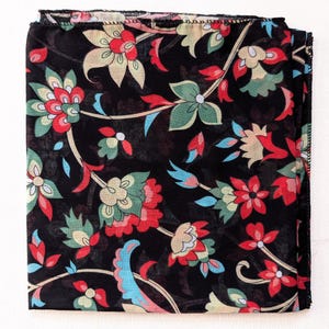 May include: Black floral scarf with a repeating pattern of red, green, and cream-colored flowers and blue accents. The scarf is square and folded, showcasing the intricate design. The edges are finished with a delicate hem.