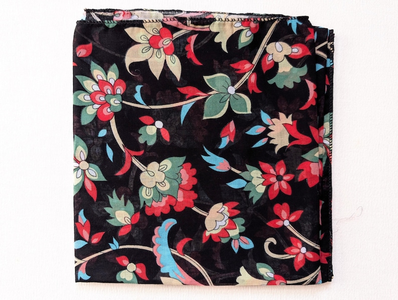 May include: Black floral scarf with a repeating pattern of red, green, and cream-colored flowers and blue accents. The scarf is square and folded, showcasing the intricate design. The edges are finished with a delicate hem.