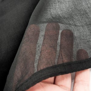 May include: Close-up of black sheer fabric. The fabric is layered, with a hand visible through the translucent material. The edges are finished with a black hem. The fabric appears lightweight and flowing.