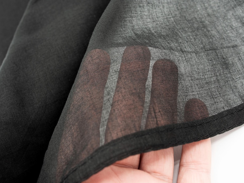 May include: Close-up of black sheer fabric. The fabric is layered, with a hand visible through the translucent material. The edges are finished with a black hem. The fabric appears lightweight and flowing.