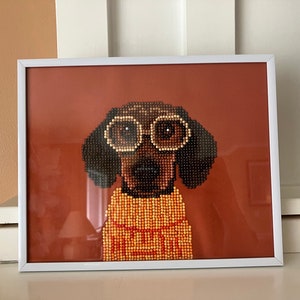 Può includere: Opera d'arte incorniciata con un bassotto pixelato con occhiali da sole e un maglione. L'immagine del cane è composta da piccole perline rotonde marroni, nere e gialle, su uno sfondo marrone-rossastro. La cornice è bianca.
