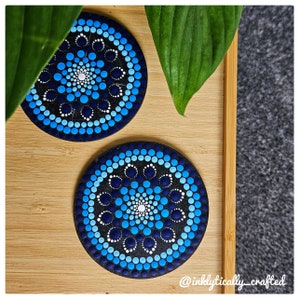 May include: Two circular coasters with a mandala design. They have a dark blue base with a pattern of blue and white dots. The coasters are on a light brown wooden tray with green leaves.