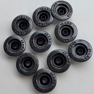 May include: A collection of ten dark gray metal jeans buttons. Each button has the word "JEANS" embossed around the outer edge. The buttons are circular with a central hole and a raised rim.