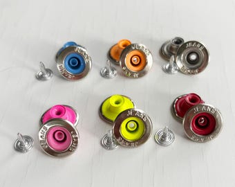 以下が含まれることがあります： 銀色の縁とマッチするリベットが付いた、カラフルなジーンズボタンのコレクション。ボタンは、青、オレンジ、ピンク、黄色、赤など、さまざまな色があります。各ボタンには「JEANS」の文字が記載されています。