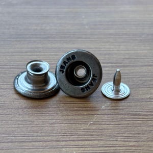 May include: Close-up of three silver-toned jean buttons. Two are assembled, and one is disassembled, showing the post and the button cap. The word "JEANS" is embossed around the edge of the button.