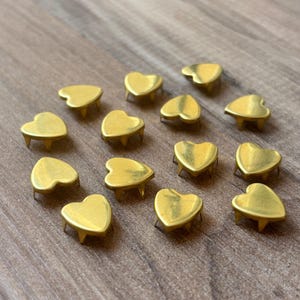 May include: A collection of heart-shaped metal studs with a gold finish. Each stud has three small legs for attachment. The studs are arranged on a wooden surface, creating a visually appealing display.