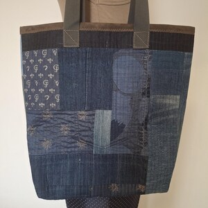 Könnte beinhalten: Eine Patchwork-Tragetasche aus verschiedenen Blautönen von Jeansstoff. Die Tasche verfügt über einen dunkelbraunen oberen Rand, graue Nylongurte und eine Mischung aus Texturen, darunter florale Stickereien und ein wiederkehrendes Lilienmuster.