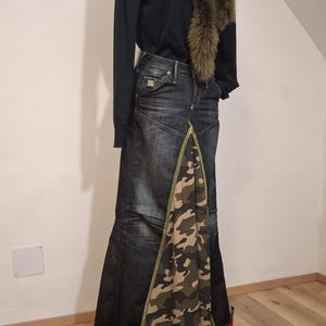 Könnte beinhalten: Ein langer dunkler Jeansrock mit einem zentralen dreieckigen Einsatz in grünem, braunem und beigem Camouflage-Muster, kombiniert mit einem schwarzen Langarmshirt, einem braunen Kunstfellkragen und Camouflage-Stiefeletten mit schwarzen Schnürsenkeln.