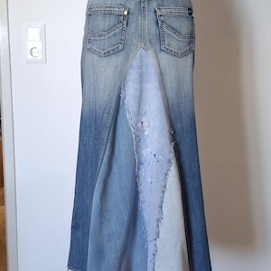 Könnte beinhalten: Ein langer Jeansrock in mittlerer Waschung mit zwei Gesäßtaschen und einem zentralen, dreieckigen Einsatz aus hellerem, abgenutztem Denim. Der Einsatz zeigt ausgefranste Kanten und weiße Farbspritzer. Ein blauer Perlengürtel wird in der Taille getragen.