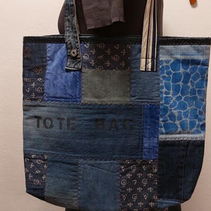 Könnte beinhalten: Eine blaue Jeans-Tragetasche mit Patchwork-Design. Die Tasche hat einen schwarzen Lederbesatz und einen weiß-schwarz gestreiften Trageriemen. Der Text "TOTE BAG" ist auf der Vorderseite der Tasche aufgedruckt.