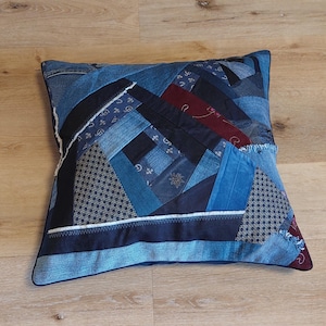 Könnte beinhalten: Quadratisches Patchwork-Kissen aus verschiedenen Blautönen von Jeansstoff. Das Kissen hat ein geometrisches Design mit verschiedenen Texturen und Mustern, darunter einfarbiger Jeansstoff, gemusterter Stoff und ein burgunderfarbener Akzent. Die Kanten sind mit einem dunkelblauen Rand versehen.