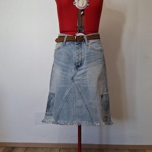 Könnte beinhalten: Hellblauer Jeansrock mit ausgefranstem Saum, präsentiert auf einer roten Schneiderpuppe. Der Rock hat einen braunen Gürtel und zwei Taschen. Die Schneiderpuppe ist rot und hat ein Singer 150-Etikett.