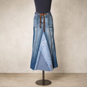 Könnte beinhalten: Ein langer Jeansrock im Patchwork-Design mit verschiedenen Blautönen, darunter ein hellblaues dreieckiges Paneel mit weißem Herzumriss, blauen Farbspritzern und einem blau-weiß gestreiften Abschnitt. Der Rock hat einen braunen Kordelgürtel.