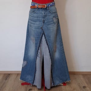 Könnte beinhalten: Jeansrock mit einzigartigem Design. Der Rock hat eine hohe Taille, einen Schlitz vorne und herzförmige Aufnäher. Er wird mit einem braunen Ledergürtel mit blau-weiß gemustertem Stoff getragen. Der Rock hat einen Used-Look.