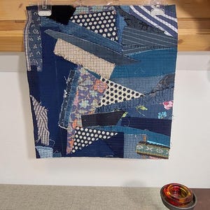 May include: A patchwork textile art piece, primarily in shades of blue, with various fabric patterns including polka dots and stripes. The artwork is hanging on a wall, and a decorative candle holder is in the foreground.