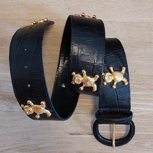 Puede incluir: Un cinturón negro con textura de relieve de cocodrilo que presenta tres adornos de ositos de metal en tono dorado sujetos a la correa. El cinturón incluye una hebilla ovalada negra a juego con un perno de metal dorado para el cierre.