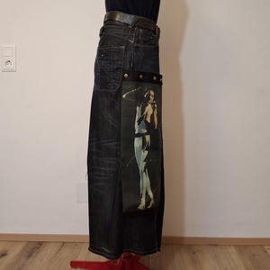 Könnte beinhalten: Dunkelblaue Jeans mit einem schwarzen Ledergürtel. Ein dekoratives Panel mit einem gedruckten Bild einer Person in weißer Kleidung ist an der Jeans befestigt. Die Jeans wird auf einer Schaufensterpuppe präsentiert.