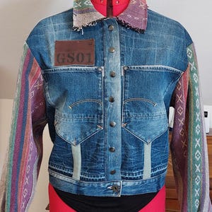 Può includere: Giacca di jeans blu con maniche e colletto fantasia colorati. La giacca presenta una toppa in pelle marrone con "GS01" sul petto. Le maniche hanno un design a righe multicolore. La giacca è esposta su un manichino.