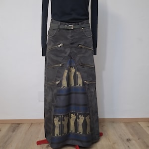 Könnte beinhalten: Langer, dunkelgrauer Rock mit mehreren Reißverschlüssen und einem dekorativen Einsatz mit goldenen Figuren auf blauem Grund. Der Rock wird mit einem schwarzen Rollkragenpullover und einem Gürtel kombiniert.