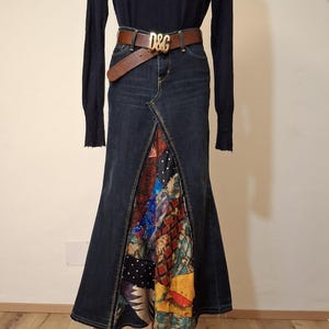 Può includere: Un dolcevita blu scuro abbinato a una gonna di jeans con un design patchwork. Una cintura in pelle marrone con fibbia dorata aggiunge un tocco elegante. L'outfit è esposto su un manichino rosso.