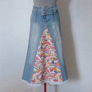 Könnte beinhalten: Ein hellblauer Jeans-Maxirock mit hoher Taille, passendem Gürtel und einer großen silberfarbenen Schnalle. Ein dreieckiger Einsatz aus weißem Stoff mit einem bunten Muster aus tropischen Blättern und Fischen ist vorne eingearbeitet und mit weißer Spitzenborte verziert.