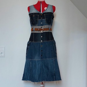 Könnte beinhalten: Ein ärmelloses Jeanskleid im Patchwork-Design mit verschiedenen Blautönen. Das Kleid verfügt über eine durchgehende Knopfleiste, zwei Brusttaschen, einen gestreiften Taillenbereich mit braunem Gürtel und einen ausgestellten Rock mit Fransen.