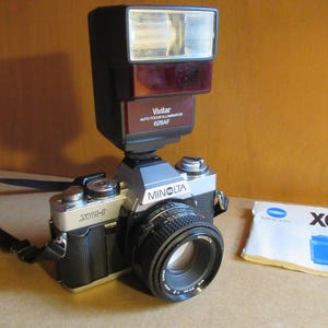 Peut inclure: Un appareil photo Minolta XG-1 vintage avec un flash Vivitar Auto Focus Illuminator 628AF fixé. L'appareil photo a un objectif noir et un corps argenté. Un petit livret d'instructions est sur le côté.