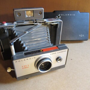 May include: Vintage Polaroid Land Camera 101 Automatic, silver and black with a red accent. The camera has a collapsible bellows and a flash attachment. The camera is shown with its black case.