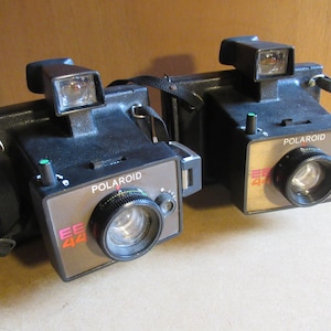以下が含まれることがあります： 2台のヴィンテージPolaroid EE44インスタントカメラ。カメラは黒色で、茶色と銀色のアクセントがあり、レンズ、ビューファインダー、ストラップが付いています。モデル名と番号はカメラの前面に印刷されています。