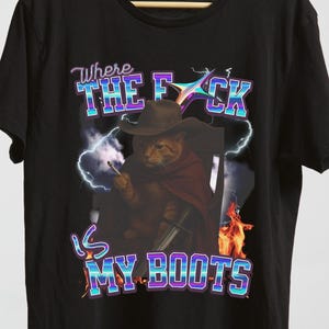 Puede incluir: Camiseta negra con un gato con sombrero de vaquero y capa, sosteniendo un cigarrillo. La camiseta muestra el texto "Where the F*ck is my Boots" en una fuente retro, con gráficos de rayos y llamas.