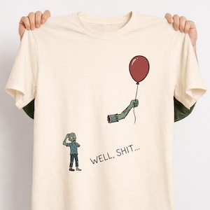 May include: A cream-coloured short-sleeve t-shirt featuring a graphic of a green zombie looking at its own detached arm floating in the air while holding a red balloon. The text "WELL, SHIT..." is printed in black below the zombie and the arm.