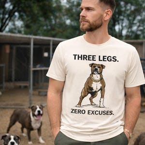 Puede incluir: Camiseta color crema con el texto "THREE LEGS. ZERO EXCUSES." presenta un perro marrón y blanco con una cadena dorada. El perro está de pie en una pose segura. La camiseta la lleva una persona.