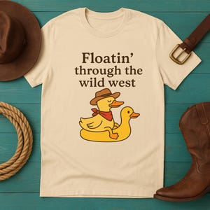 Peut inclure: T-shirt crème avec l'inscription "Floatin' through the wild west". Le t-shirt présente un canard de dessin animé portant un chapeau de cow-boy et un bandana rouge sur un canard gonflable jaune. Une botte et un chapeau de cow-boy bruns sont visibles.