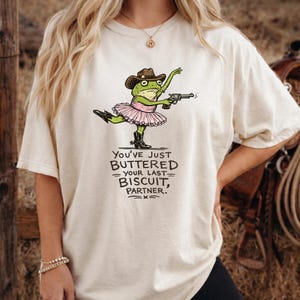 Puede incluir: Camiseta color crema con una rana de dibujos animados con sombrero de vaquero, tutú y botas, sosteniendo una pistola. El texto "You've just buttered your last biscuit, partner" está impreso debajo.