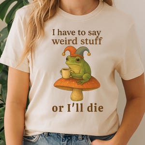 Op de afbeelding: Crèmekleurig T-shirt met een grillige illustratie van een groene kikker met een narrenmuts en een mok, zittend op een paddenstoel. De tekst "I have to say weird stuff or I'll die" is in bruin gedrukt.
