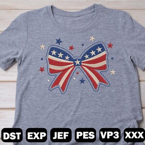May include: A gray t-shirt featuring an embroidered bow design in red, white, and blue, with stars. The bow is the central focus, surrounded by additional star embellishments. The bottom of the shirt displays the text "DST EXP JEF PES VP3 XXX".