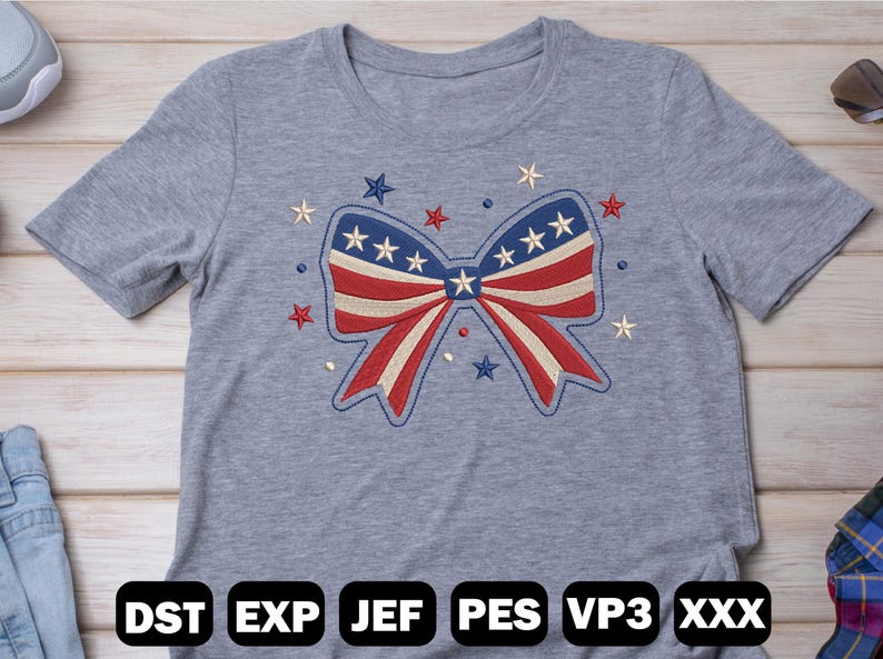 May include: A gray t-shirt featuring an embroidered bow design in red, white, and blue, with stars. The bow is the central focus, surrounded by additional star embellishments. The bottom of the shirt displays the text "DST EXP JEF PES VP3 XXX".