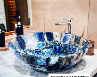 May include: A blue and white agate stone vessel sink. The sink is oval-shaped and features a mosaic pattern of blue, white, and gray stones. The sink is sitting on a white countertop. The text "EpoxyMarbleAgateShop" is at the bottom.