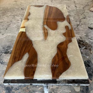 May include: A rectangular wooden table with a unique design. The table features sections of dark brown wood and creamy white epoxy resin. The surface is smooth and glossy, with the text "EpoxyMarbleAgateShop" visible.