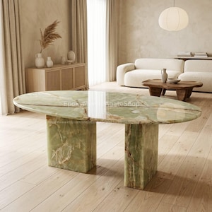 May include: An oval-shaped green onyx table with a polished surface. The table is supported by two rectangular bases. The room has light-colored walls and wooden floors. The table's dimensions are not visible.