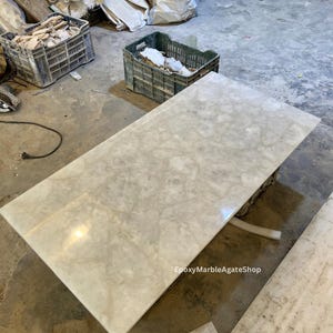 May include: A large, white, polished slab of marble with a veined pattern. The marble is sitting on a concrete floor. The text "EpoxyMarbleAgateShop" is visible in the lower right corner of the image.