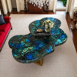 Könnte beinhalten: Einzigartiger Couchtisch in freier Form mit einer schillernden blau-grünen Oberfläche. Der Tisch steht auf einem goldfarbenen Gestell und ist mit einem Blumenarrangement versehen. Maße sind nicht sichtbar.